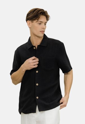 Ung mann med stylet brunt hår iført en svart kortermet skjorte med knapper og hvite bukser, ser til siden mens han justerer en knapp.