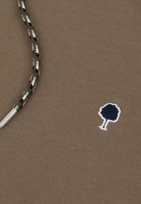 Tessuto marrone con una trama texturizzata, presenta un piccolo ricamo di un albero blu navy e un cordone decorato con accenti verdi, neri e bianchi.