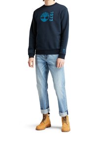 Sudadera de marina con logo turquesa y texto "1973", combinada con jeans claros arremangados y botas amarillas con cordones marrones.