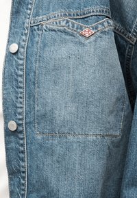 Nahaufnahme einer Tasche aus blauem Denim mit schräger Naht und kleinem roten "Replay"-Logo-Pin am oberen Rand.