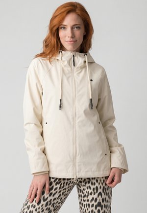 Frau mit roten Haaren, die eine hellbeige, mit Reißverschluss versehene Jacke mit Kordelzügen und Leopardenmuster-Hose trägt, vor einem schlichten Hintergrund steht.