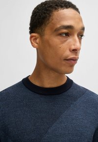 Maglione lavorato a maglia con un motivo geometrico blu navy e azzurro, caratterizzato da un collo rotondo a coste nere e un tessuto testurizzato.