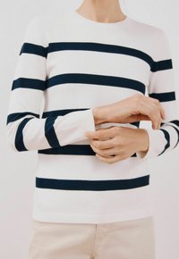 Maglione bianco con righe orizzontali blu navy, collo rotondo, polsini e orlo a coste, design aderente, indossato su un braccio con mani che regolano la manica.