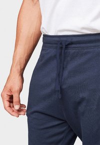 Un bras et une main masculine à côté de pantalons de survêtement bleu marine avec cordon de serrage, associés à un t-shirt blanc rentré, contre un fond clair uni.