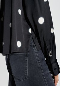 Black blouse with white polka dots, featuring long sleeves and button cuffs, paired with dark gray denim jeans. Smooth fabric with subtle sheen.