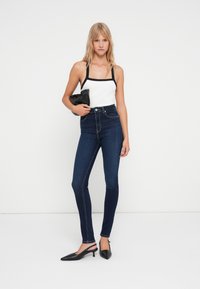 Tamne traperice visokog struka tamne boje u kombinaciji s bijelim topom bez rukava s crnim detaljima. Model drži crnu clutch torbicu i nosi crne cipele s visokim potpeticama.