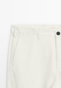 Pantalon en velours côtelé blanc avec une taille plate, des poches latérales et un détail de bouton noir à l'avant. Le tissu a une texture côtelée.