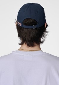Marineblaues Basecap aus Baumwolle, mit einem verstellbaren Riemen aus Metall hinten und einem pinken Logo-Patch auf der Rückseite.