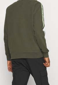 Sudadera verde oliva con puños y dobladillo acanalados, con un detalle de cinta con logo en las mangas. Tejido suave y ajuste relajado.