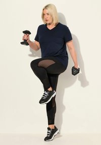 Granatowa koszulka treningowa z dekoltem w serek, czarne legginsy z siateczkowymi wstawkami oraz czarne buty sportowe z białymi sznurówkami. Trzyma hantle w każdej ręce.