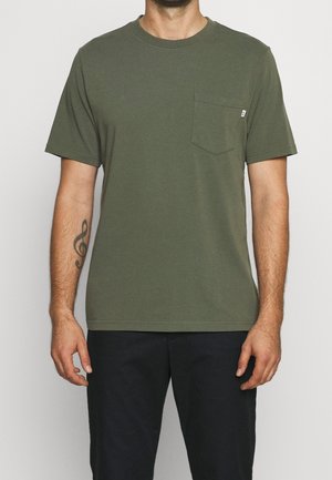 Mężczyzna w oliwkowozielonym t-shircie z krótkim rękawem i kieszenią na piersi oraz czarnych spodniach, stojący z rękami swobodnie opartymi po bokach.