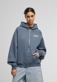 Plava dukserica s patentnim zatvaračem, s kangaroo džepom, rebrastim manžetnama i žutim logotipom na prsima. Mekani materijal s opuštenim krojem i kapuljačom.