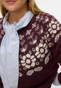 Pull en maille bordeaux avec un motif floral crème et des accents roses, associé à un blouse bleu clair avec un nœud au cou. Texture douce et coupe décontractée.