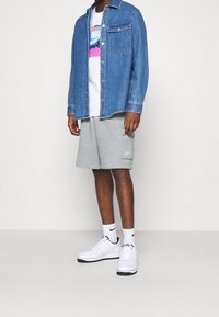 Man som bär en blå jeansjacka, vit Nike t-shirt med grafiskt tryck, grå Nike shorts, vita Nike strumpor och vita Nike sneakers.