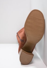 Bottine en daim marron avec une fermeture éclair latérale, une semelle inclinée montrant les rainures, et un petit talon bloc sur fond blanc.