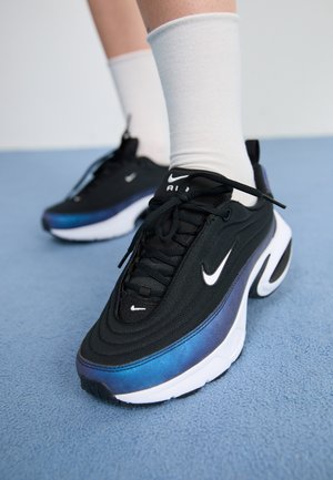 Personne portant des baskets Nike Air Max noires et bleues avec des chaussettes blanches, debout sur un sol bleu texturé.
