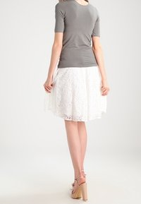 Graues kurzärmeliges Oberteil kombiniert mit einem weißen Spitzenrock mit floralen Mustern. Das Model trägt nude-farbene Sandalen mit Blockabsatz.