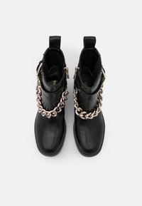 Bottes basses en cuir noir avec détail de chaîne dorée sur le devant et fermetures éclair latérales, vues de dessus sur fond blanc.