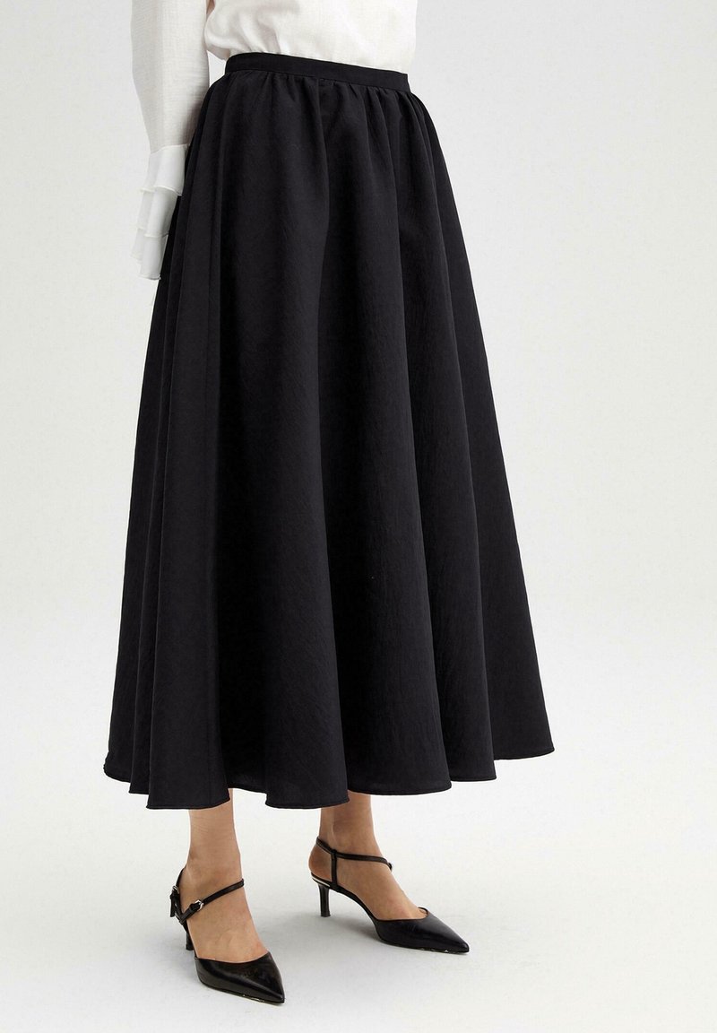 Jupe noire taille haute, longueur totale, portée avec des talons noirs à bride cheville à bout pointu sur fond blanc.