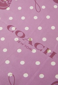 Fialová látka s bielym bodkovaným vzorom, s textom "COACH NEW YORK" vytlačeným tmavšou fialovou farbou. Hladký povrch.
