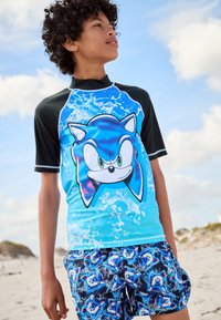 Kortærmet rashguard i blå med sorte detaljer, med et Sonic-karakterdesign og bølgemønstre. Badeshorts med matchende Sonic-print.