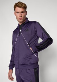 Lilla hettejakke med svarte detaljer, med høy krage, hvit piping og tre svarte striper på ermene. Laget av glatt stoff.