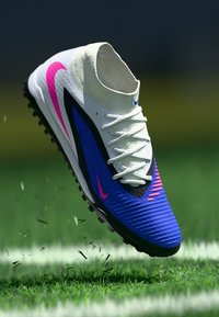 Ποδοσφαιρικό παπούτσι Nike με λευκό πάνω μέρος, μπλε και ροζ λεπτομέρειες, υφή σχεδίασης και παραδοσιακό δέσιμο κορδονιών, αιωρούμενο πάνω από χορτάρι.