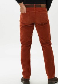 Uomo che indossa pantaloni di corduroy color ruggine con una cintura marrone e scarpe marrone scuro, mano nella tasca posteriore, in piedi su uno sfondo bianco.