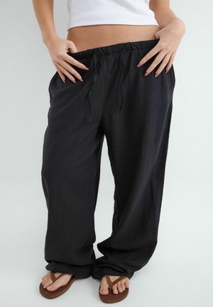 Osoba nosiaca voľné čierne nohavice a hnedé žabky, drží si pás nohavíc, s odhaleným bruchom a bielym topom.