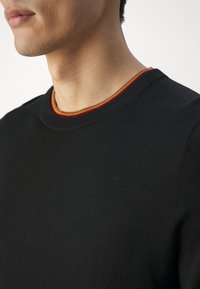Gros plan sur une personne portant un pull noir à col rond avec une bordure rayée rouge, jaune et orange le long du col.