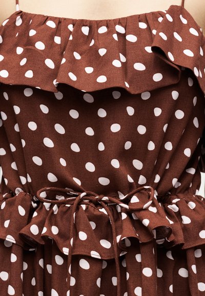 Καφέ αμάνικο φόρεμα με λευκές πουά, βολάν στο ντεκολτέ και στη μέση, δεμένο με ζώνη από το ίδιο ύφασμα.