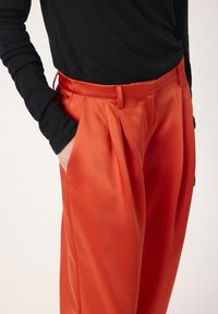 Pantalons en satin orange avec une texture lisse, dotés de plis à la taille et de poches latérales. Associés à un haut noir à manches longues.