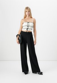 Donna bionda che indossa un top corto bianco a ruches e pantaloni neri larghi a vita alta, con una borsa nera, in piedi contro uno sfondo bianco.