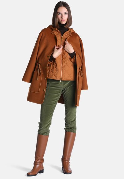 Jeune femme portant un manteau cape marron par-dessus une veste matelassée, un pantalon vert et de longues bottes marron, debout devant un fond uni.