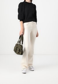 Pull noir avec des manches bouffantes texturées, jean évasé crème, et sac à main en tissu vert avec plusieurs poches, porté avec des baskets blanches.