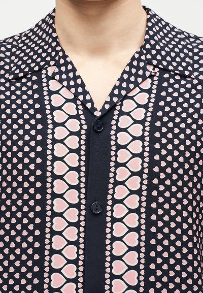 Κοντινό πλάνο ενός ναυτικού μπλε πουκάμισου με ροζ μοτίβα καρδιάς διατεταγμένα σε κάθετες ρίγες και δύο μαύρα κουμπιά στον εμπρόσθιο καβάλο.