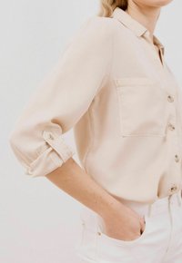 Camicia beige con bottoni e maniche arrotolate, dotata di una tasca frontale e tessuto morbido. Il modello indossa pantaloni bianchi, mostrando una vestibilità rilassata.