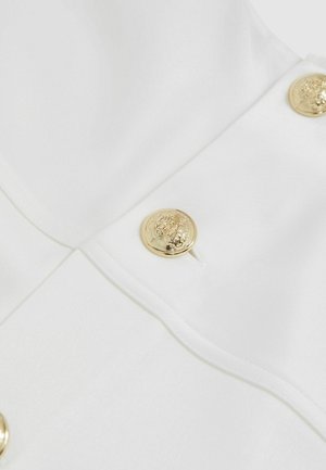 Balts audums ar gludu tekstūru, rotāts ar dekoratīvām zelta pogām ar sarežģītu dizainu aizdarei.