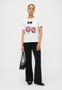 T-shirt en coton blanc avec un imprimé graphique de cerise et de nœud, associé à un pantalon noir évasé et un petit sac à main noir. Tenue simple et décontractée.