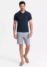 Marinblå polotröja med korta ärmar, ljusgrå shorts och beige mockaskor. Har en diskret logotyp på polotröjans bröst.