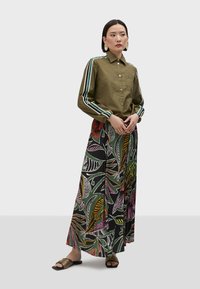 Olive green button-up shirt with striped sleeves, paired with a long, black skirt featuring colorful leaf patterns and flat sandals.