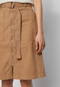 Ruskea puuvillahame, jossa on edessä vetoketju, kaksi suurta taskua ja leveät vyölenkit. Tekstuuri on sileä, ja helman reunassa on ompeluyksityiskohtia.