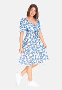 Vestido floral con estampados azules, blancos y rojos, cintura ajustada, mangas abullonadas, falda a la altura de la rodilla, combinado con zapatillas blancas.
