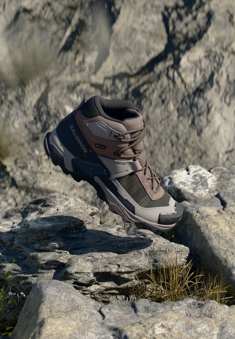 Botas de caminhada castanhas e cinzentas com uma superfície texturizada, sistema de atacadores e sola de borracha, posicionadas em terreno rochoso, mostrando um design robusto.