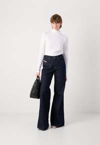 Dark blue denim flared jeans with fitted waist, contrasted stitching, and side pockets. Paired with a white long-sleeve turtleneck top and black handbag.