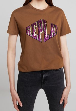 Kvinde iført en brun T-shirt med lilla og guld paillet-tekst "REPLAY", kombineret med sorte jeans mod en ensfarvet grå baggrund.