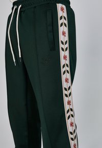 Pantalones de chándal verdes con cordón, que cuentan con bolsillos laterales y un patrón de rayas blancas con rosas rojas y hojas verdes. Logo bordado en el bolsillo.
