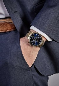 Montre chronographe noir et bleu avec un bracelet en métal doré, un cadran rond, des chiffres en gros caractères et des sous-cadrans, portée sur un poignet dans une veste de costume.