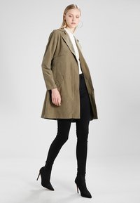 Mujer rubia caminando con un abrigo verde oliva hasta las rodillas, camiseta blanca, pantalones skinnies negros, botas de tacón alto negras hasta el tobillo y grandes pendientes geométricos.