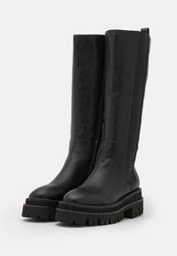 Botas de cuero negras hasta la rodilla con paneles elásticos laterales, suela de goma gruesa y detalles de tracción texturizada en la base.